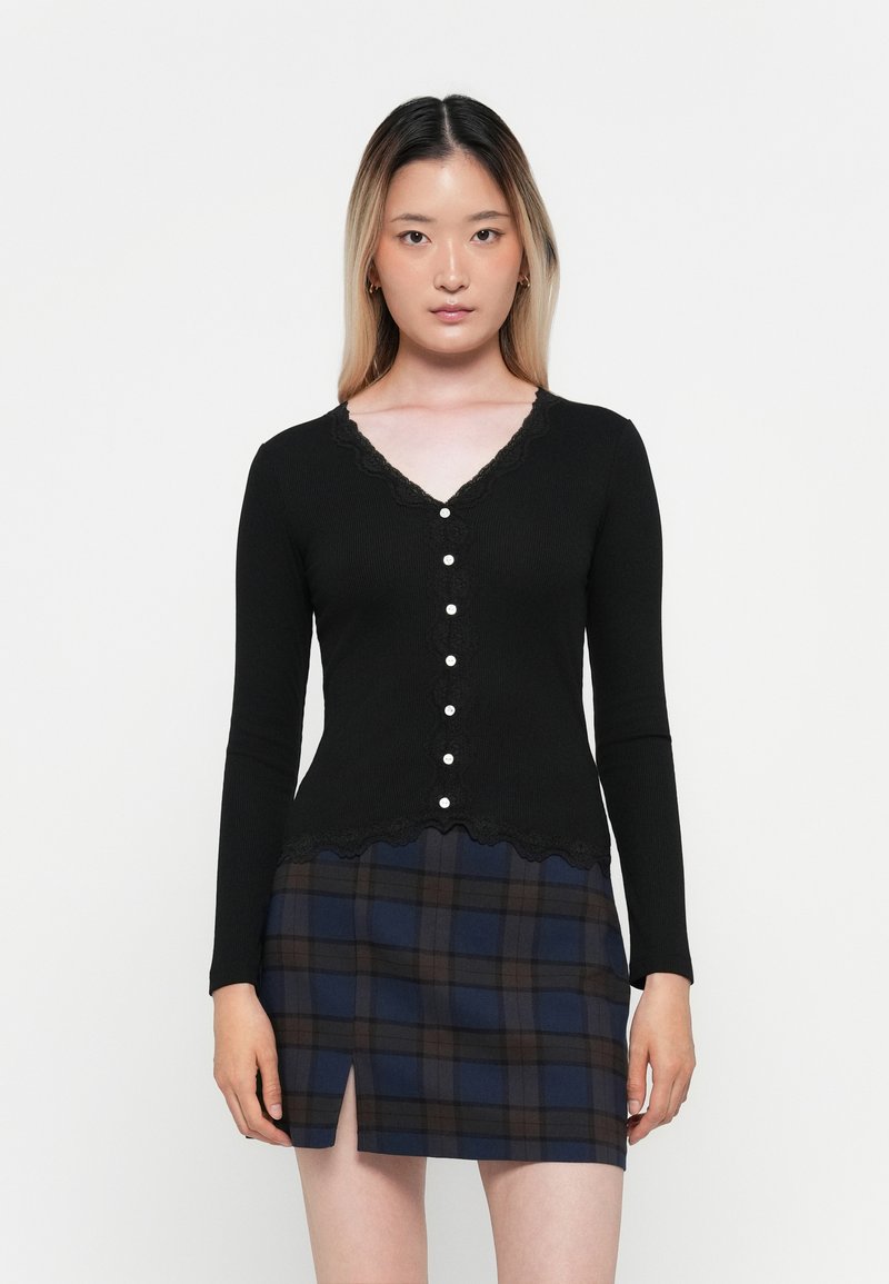 Cardigan noir à côtes avec des manches longues, col en V, bordure en dentelle festonnée, fermeture à boutons, associé à une mini-jupe à carreaux dans des tons de bleu foncé et de marron.