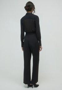 Femme avec les cheveux en chignon portant un blouse noire transparente à manches longues, un pantalon noir taille haute et des escarpins noirs, devant un fond uni.