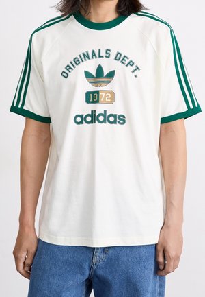 Persoon draagt een wit Adidas Originals Dept. t-shirt met groene bies en retro logo, gecombineerd met blauwe jeans, staande tegen een witte achtergrond.