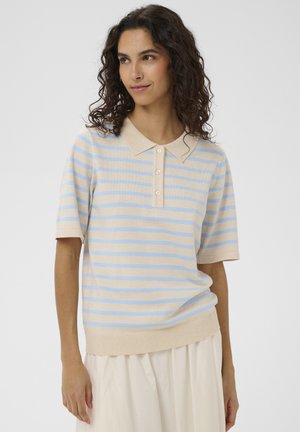 Žena s kučeravými tmavými vlasmi, oblečená v béžovo-svetlomodrom pruhovanom krátkom rukáve polo trička a svetlej sukni, stojaca pred bielym pozadím.