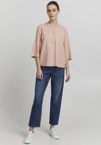 Blouse en lin rose avec un col henley à boutons, des manches trois-quarts et une coupe décontractée, associée à un jean bleu à jambes droites et des baskets blanches.