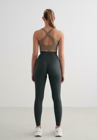 Sostenitore sportivo marrone con spalline incrociate e schiena a taglio, abbinato a leggings verdi scuri a vita alta. Texture liscia e design aderente.