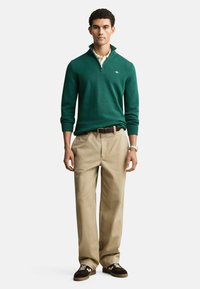 Grüner Pullover mit Reißverschlusskragen, kombiniert mit beigen Hosen und braunen Sneakers. Der Pullover hat gerippte Bündchen und ein Logodetail auf der Brust.