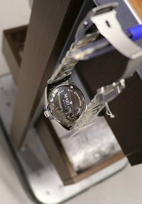 Montre en acier inoxydable argenté avec un bracelet en cuir noir, cadran rond, fond en verre clair et détails gravés sur le boîtier arrière.