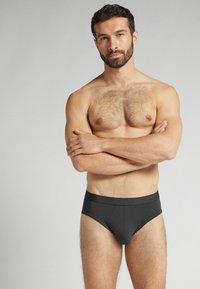 Schwarze Boxershorts mit glatter Textur und einem auffälligen Bund, Modell steht mit verschränkten Armen und zeigt ein sauberes, minimalistisches Design.