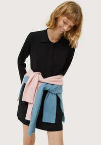 Vestido polo negro de manga larga con botonera; suéteres de punto en rosa y azul atados en la cintura, añadiendo textura y contraste de color.