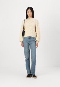 Maglione a maglia giallo chiaro con un motivo tessile, abbinato a jeans blu chiaro a gamba dritta e sneaker nere. Piccola borsa nera appesa alla spalla.