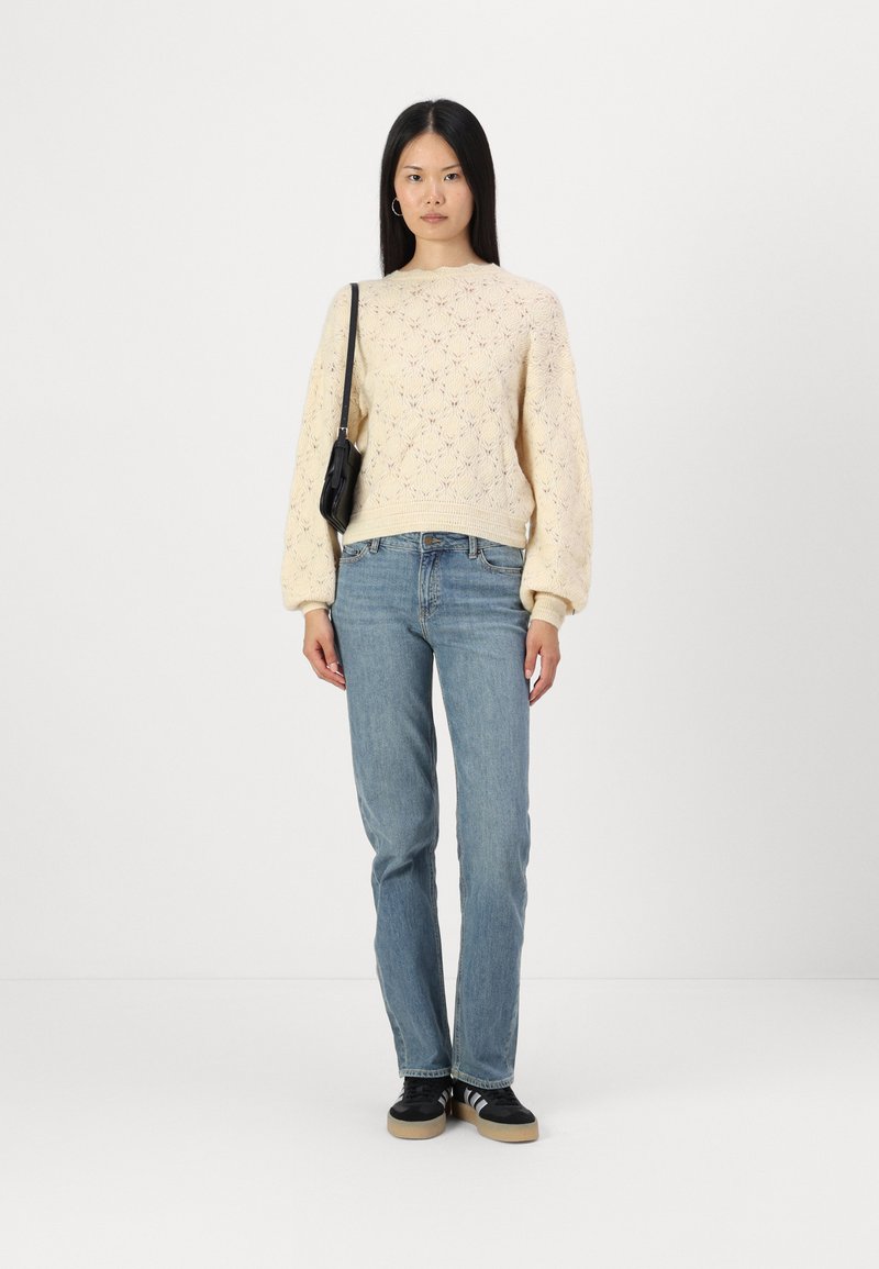 Maglione a maglia giallo chiaro con un motivo tessile, abbinato a jeans blu chiaro a gamba dritta e sneaker nere. Piccola borsa nera appesa alla spalla.