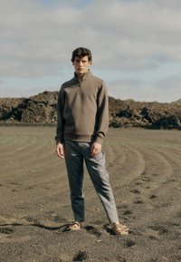 Sweatshirt z zipom v olivno zeleni barvi, v kombinaciji s svetlo sivo teksturiranimi hlačami in bež sandali, postavljenimi v skalnatem okolju.