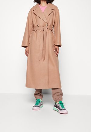 Lange beige wollen blend jas met een ceintuur in de taille, dubbele knopen, brede revers en lange mouwen. Gecombineerd met lichtroze en groene sneakers.