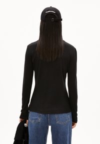 Schwarzes, langärmliges Oberteil mit taillierter Passform, getragen mit hochgeschnittenen blauen Jeans. Auf dem Kopf trägt die Person eine schwarze Mütze mit weißem Schriftzug.