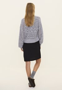 Hellgrauer, gestrickter Pullover mit gemustertem Textur, kombiniert mit einem schwarzen, figurbetonten Kleid, grauen Socken und dunklen Slippern.