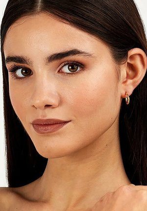 Goldcreolen an einem Model mit glattem, dunklem Haar, samtiger Haut und neutralem Make-up, das die natürlichen Merkmale und Gesichtskonturen betont.