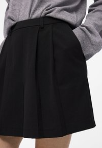 Jupe plissée noire en tissu texturé, avec des poches latérales et une taille élastique. Le haut est gris avec une silhouette ajustée.