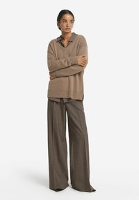 Beige Strickpullover mit V-Ausschnitt, darüber mit einem braunen Hemd mit Kragen kombiniert, dazu weite braune Hose mit strukturiertem Finish.