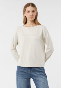 Béžová košile s dlouhým rukávem a kulatým výstřihem, s nápisem "MINIMALIST" v jemném, reflexním materiálu. Měkká látka s volným střihem.