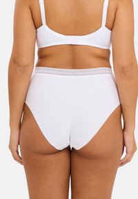 Femme portant une lingerie blanche taille haute et un soutien-gorge blanc, vue de dos contre un fond clair.