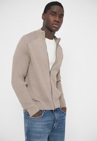 Béžový svetor na zips s rebrovanou textúrou, vysokým golierom a priliehavými manžetami. Nosený cez bielu košeľu, doplnený modrými džínsy.