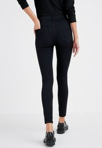 Jean skinny noir taille haute avec une texture lisse, doté de deux poches arrière et d'une coupe slim. Porté avec des chaussures noires.