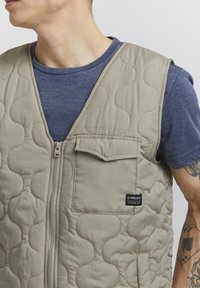 Beige quilted vest with a zipper closure, featuring a front pocket and wavy stitching pattern. Worn over a navy blue shirt.