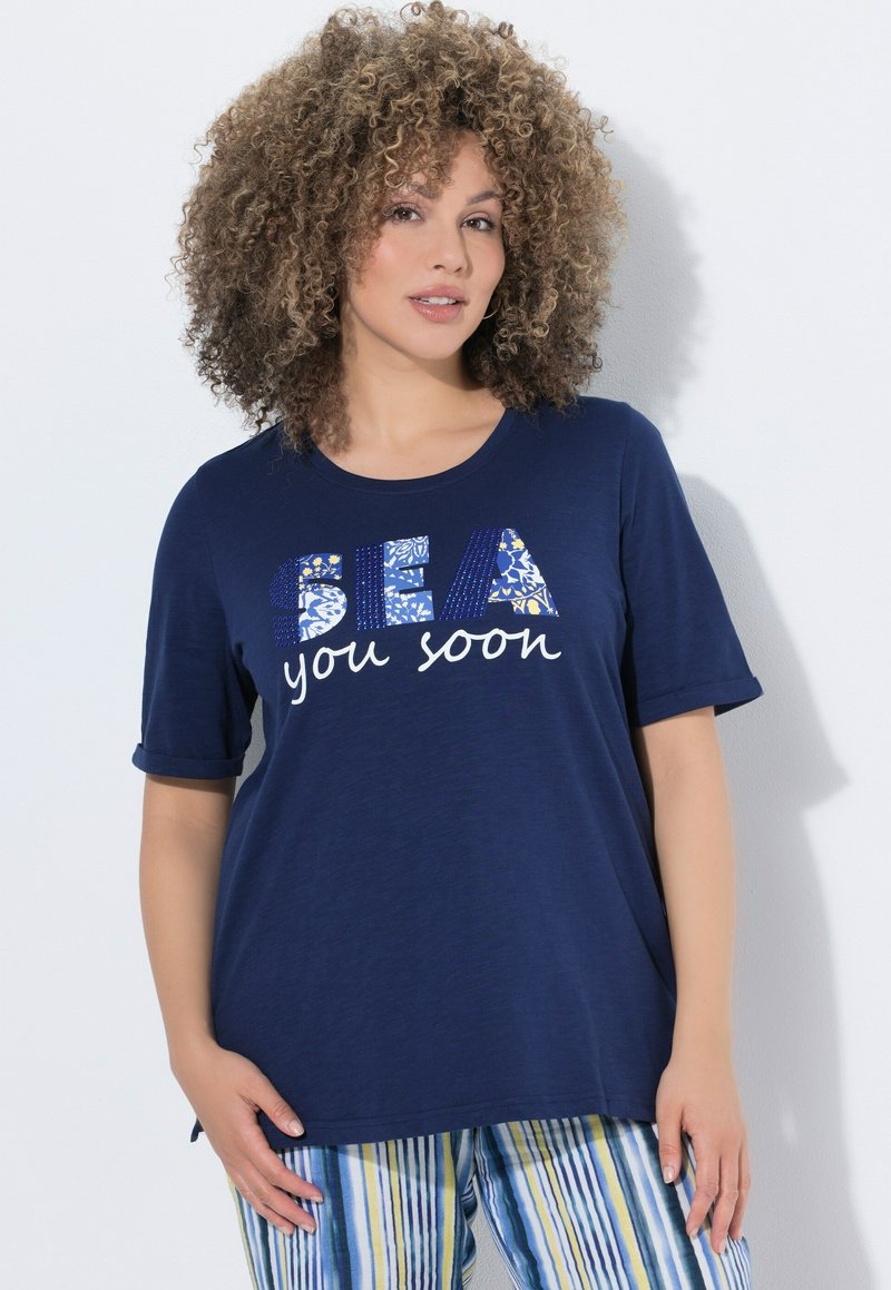 Frau mit lockigem Haar, die ein marineblaues Hemd mit dem Aufdruck "SEA you soon" und blau-gelb-weiß gestreifte Hosen trägt, vor einem weißen Hintergrund stehend.