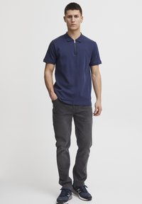 Polo de color azul marino con cuello con cremallera, combinado con jeans grises y zapatillas azul marino. La camisa presenta un logo sutil en el pecho.