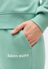 Χέρι που ακουμπά στην τσέπη ανοιχτοπράσινου παντελονιού φόρμας Björn Borg, δείχνοντας την υφή του υφάσματος και τη λεπτομέρεια του λογότυπου.