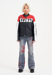 Black leather jacket with red and white color blocks, zipper closure, and logo patches; paired with gray flared jeans featuring red embroidery.