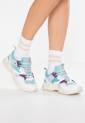 Piernas con calcetines blancos hasta la mitad de la pantorrilla con rayas beige y zapatillas deportivas gruesas en blanco, turquesa y morado con suelas beige gruesas.