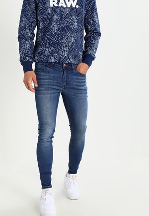 Osoba ubrana w niebieską koszulę z długim rękawem w wzory z napisem „RAW.”, dopasowane niebieskie dżinsy i białe trampki, stojąca z jedną ręką w kieszeni.