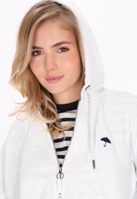 Felpa zip grigia chiara con tasca frontale, cappuccio con coulisse e un logo di ombrello ricamato in blu scuro sul petto. Maglietta a righe sotto.