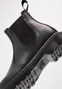 Bottine Chelsea en cuir noir avec panneau latéral élastique, boucle à l’arrière et semelle épaisse crantée inclinée vers le haut.