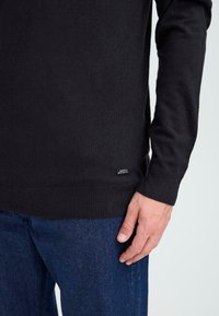 Pull en tricot noir à manches longues, avec des poignets et un ourlet côtelés, arborant une petite étiquette sur le côté inférieur, associé à un pantalon en jean bleu foncé.