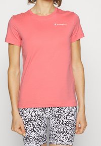 T-shirt in cotone corallo con maniche corte e colletto girocollo, con un logo Champion bianco sul petto. Abbinato a pantaloni corti a fantasia bianca e nera.