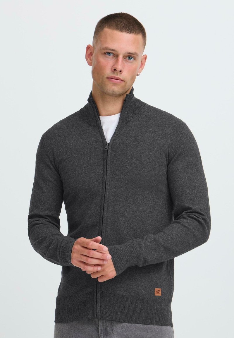 Hombre vistiendo un suéter gris oscuro con cremallera sobre una camisa blanca, de pie con las manos entrelazadas contra un fondo claro y sencillo.