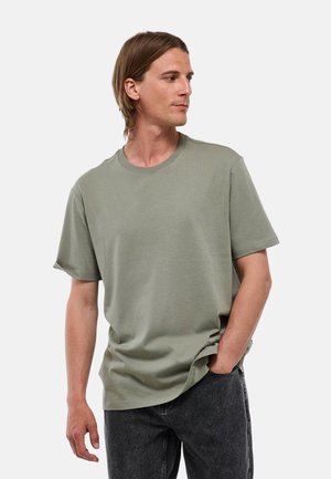 Homme aux cheveux bruns mi-longs portant un t-shirt vert clair à manches courtes et un jean gris foncé, regardant vers sa droite avec une main dans la poche.
