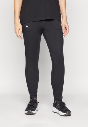 Persoon draagt zwarte Under Armour leggings en zwarte sportschoenen met witte zolen, staand tegen een effen achtergrond.
