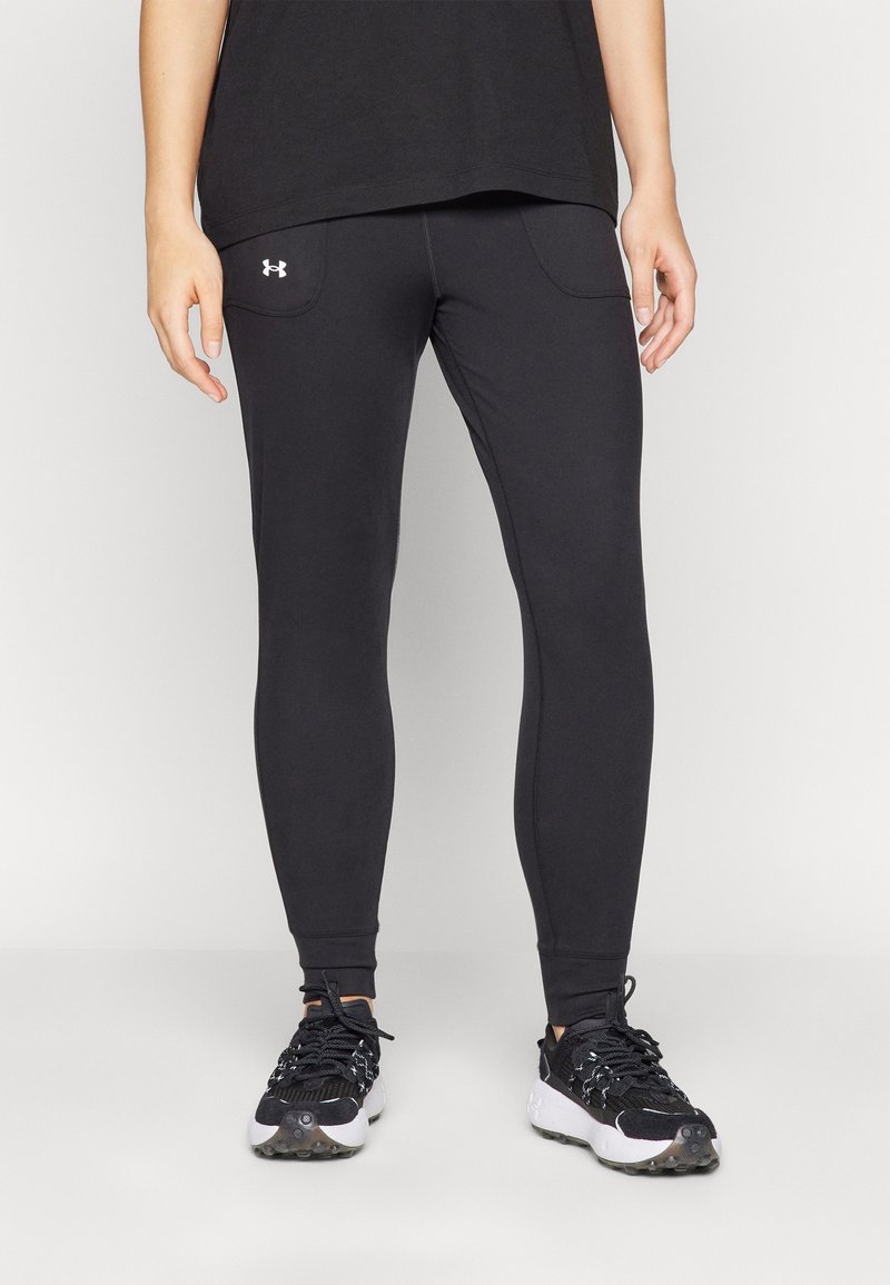 Personne portant un legging noir Under Armour et des chaussures de sport noires avec des semelles blanches, debout devant un fond uni.