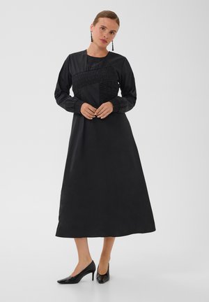 Femme portant une robe midi noire à manches longues avec détails froncés, des talons noirs et des boucles d’oreilles pendantes, debout devant un fond blanc uni.