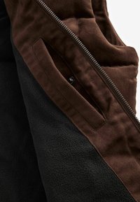 Brun og sort jakke med lynlås, glat stoffblanding og en sidelomme med knap. Tekstureret yderlag og blødt indre fleece.