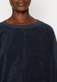 Top de tejido suave en color azul marino, con un escote ancho y un ajuste holgado. Presenta una costura mínima y un diseño sin costuras para una apariencia suave.