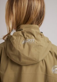 Giacca cappuccio khaki con una texture morbida, caratterizzata da un logo e coordinate stampate sul retro. Il cappuccio è dotato di un cordino per la regolazione.
