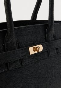 Détail d'un sac à main en cuir noir texturé avec deux anses et une boucle rectangulaire dorée à morsure tournante sur la sangle avant.