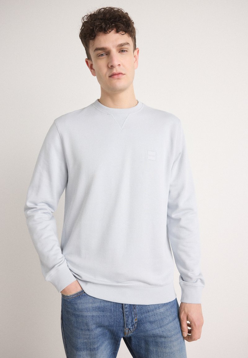Sudadera azul clara con cuello redondo, mangas largas, puños y dobladillo acanalados, que presenta un pequeño logo en relieve en el pecho. Combinada con jeans.