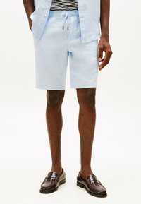 Shorts in lino azzurro chiaro con vita elastica e dettaglio con cordino, abbinati a mocassini in pelle marrone. Texture liscia, lunghezza fino a metà coscia.