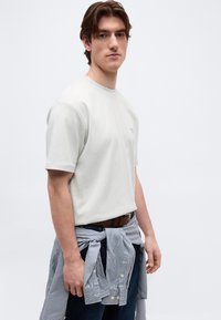 Νέος άνδρας που φοράει λευκό κοντομάνικο μπλουζάκι, ριγέ πουκάμισο δεμένο γύρω από τη μέση και σκούρο παντελόνι, στέκεται μπροστά σε απλό φόντο.