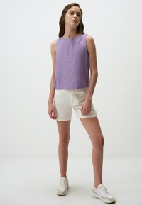 Lavendel mouwloze top gemaakt van lichte stof, gecombineerd met witte denim shorts. Model draagt witte sneakers met beige accenten op een effen achtergrond.