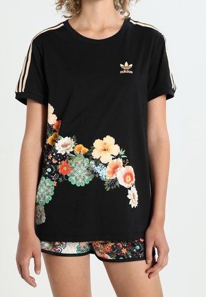 T-shirt noir Adidas avec des rayures beiges sur les manches et un imprimé floral sur le devant, porté avec un short à motifs.
