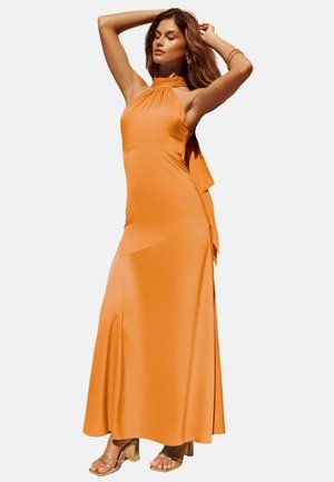 Frau posiert mit geschlossenen Augen, trägt ein ärmelloses orangefarbenes Maxikleid mit Neckholder und goldene Sandalen mit hohen Absätzen vor weißem Hintergrund.