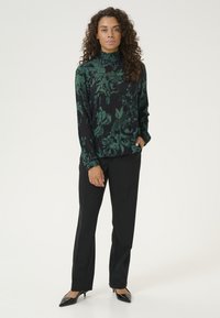 Blusa nera a fiori con collo alto e maniche lunghe, caratterizzata da un motivo floreale verde; abbinata a pantaloni neri su misura e tacchi neri.